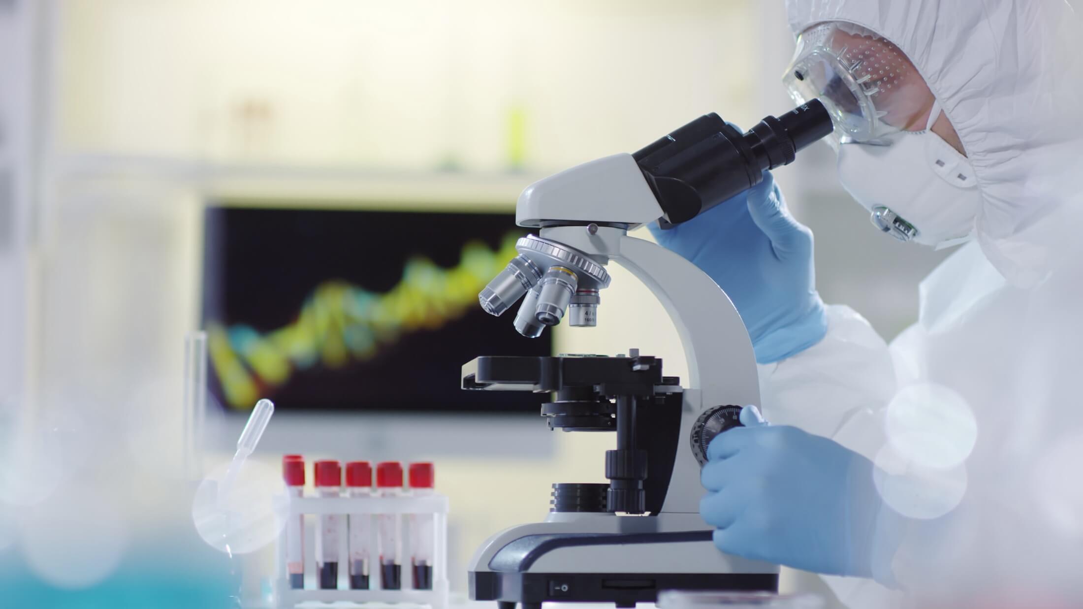 Lab Technician in protective gear is looking through a microscope.